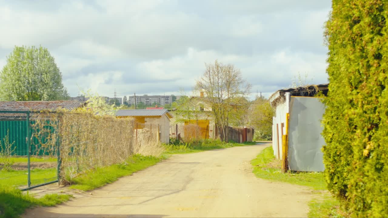 Dirt road lined with sheds and trees under a partly cloudy sky, with urban apartment blocks in the distant background. Daugavpils, Latvia (Latgale)