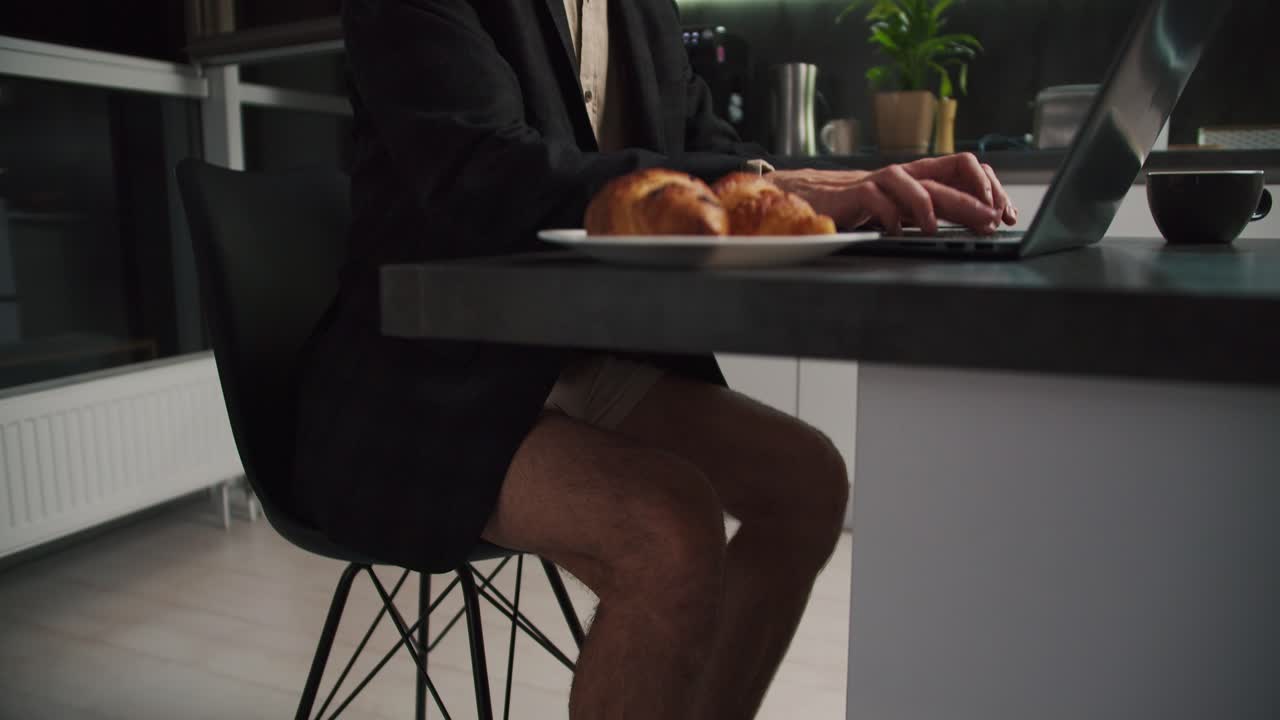 un hombre moreno con paja en una chaqueta pero sin pantalones de oficina se sienta frente a su portátil y trabaja mientras se comunica a través de videoconferencia en una cocina moderna por la noche