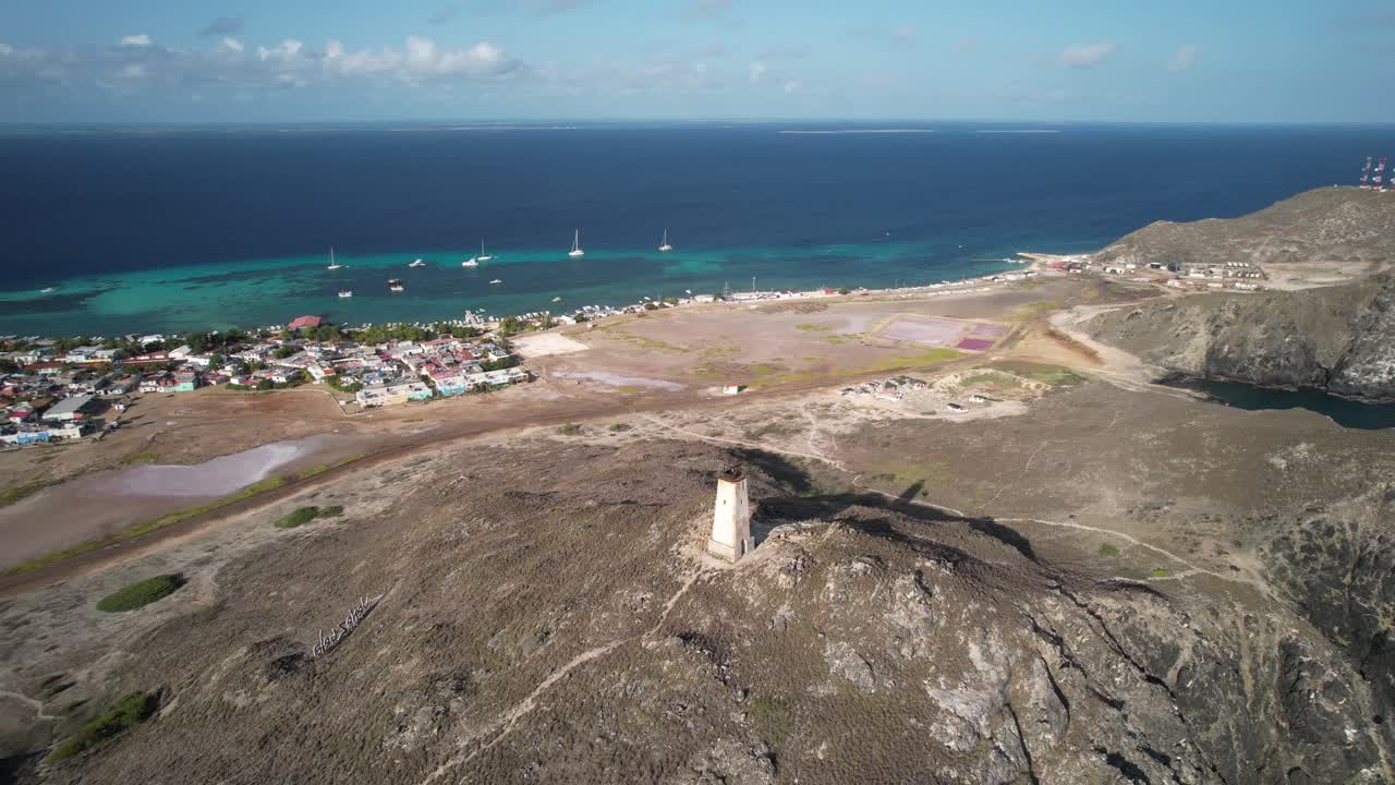 Gran roque island with a lighthouse on a hill and vibrant blue waters in the distance, aerial view