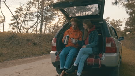 young couple sitting in the open trunk of the car having a hot drink with a thermos