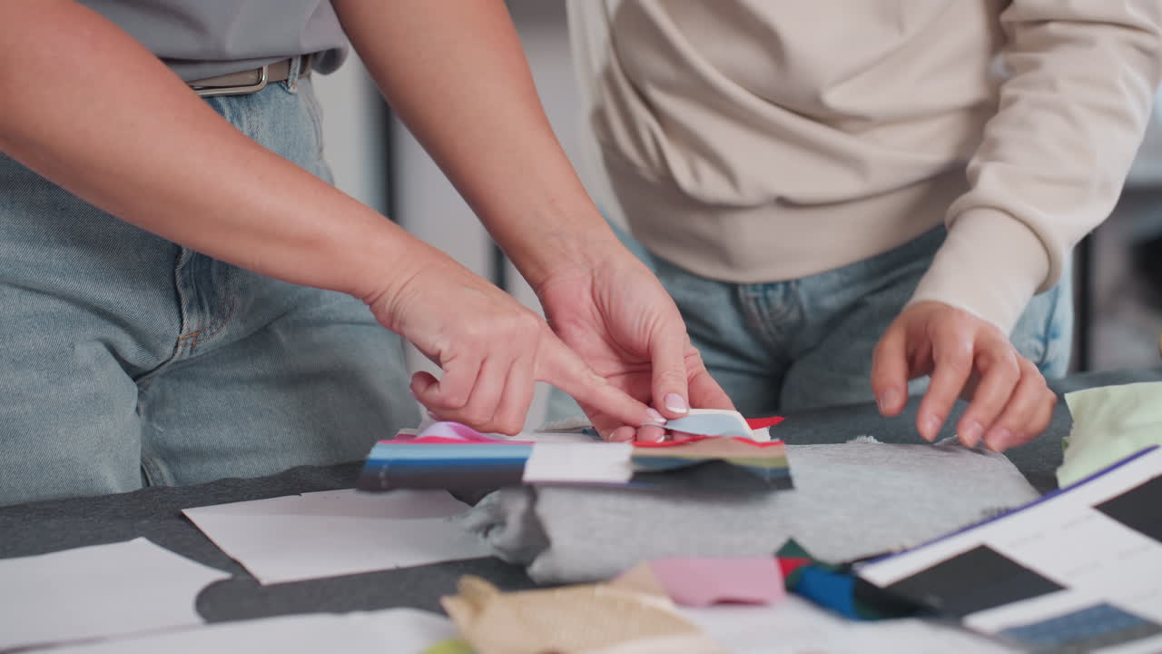 Fabric designers hand view comparing textile color samples at creative workspace focusing on matching shades for fashion project surrounded by colorful fabric swatches notebooks sketches