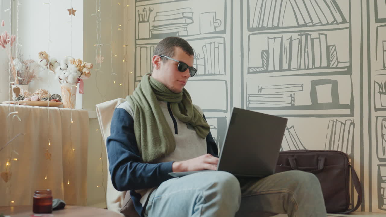 White man with laptop in study, scarf and sunglasses, typing quietly by window with warm light, bookshelf mural backdrop, coffee and vase on side table, relaxed focused freelance writer creating