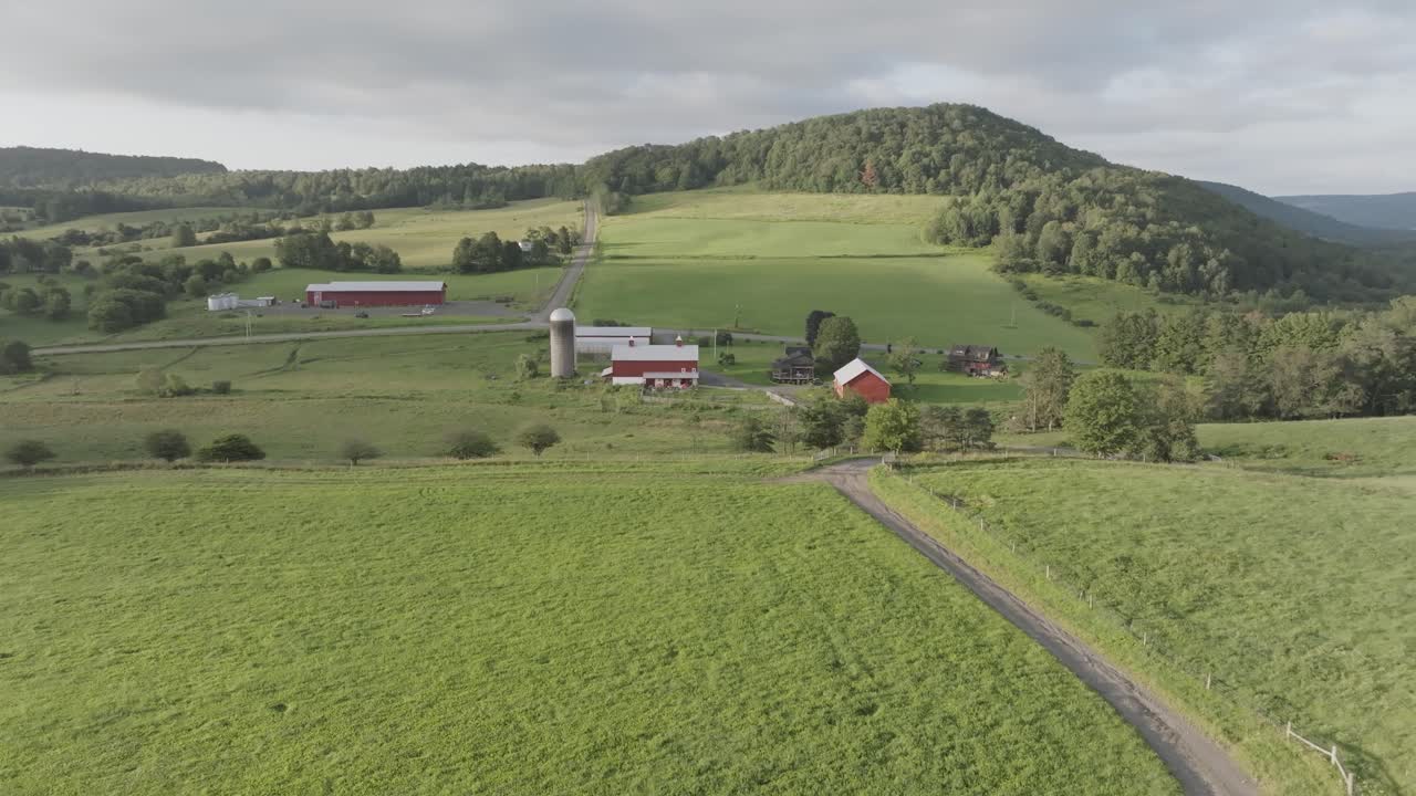 Rolling green fields surround a farm in Upstate New York, with red barns, a silo, and a winding country road leading toward a forested hillside.
