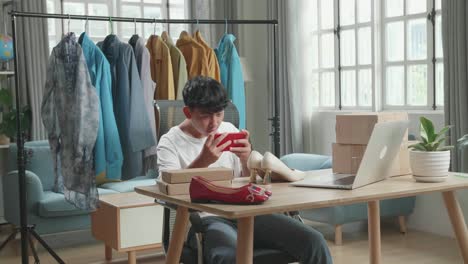 asian man online seller taking photos with mobile phone while using computer for selling clothes at home