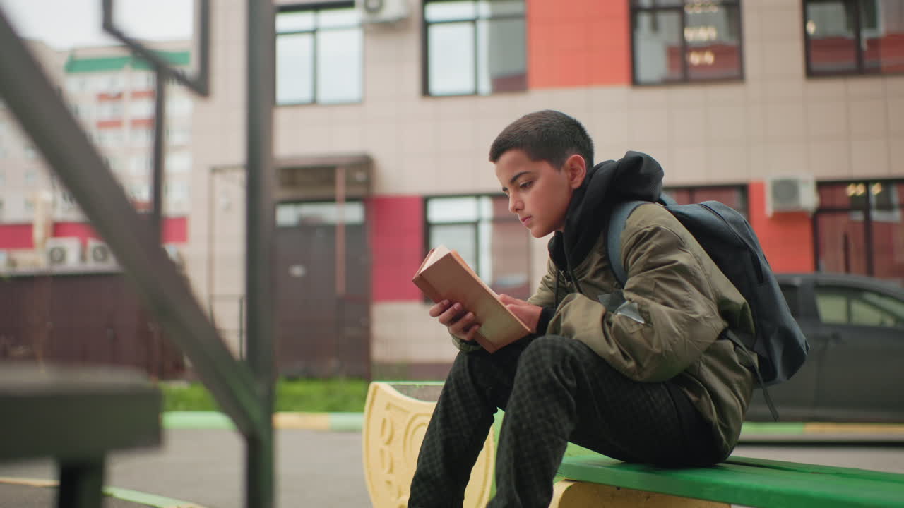 Side view of young boy in green jacket and backpack flipping through pages of novel while seated outdoors with blurred city building in background