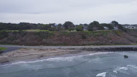 Aerial-orbit-crashing-waves-on-beach-with-drift-wood,-amazing-ocean-view-homes