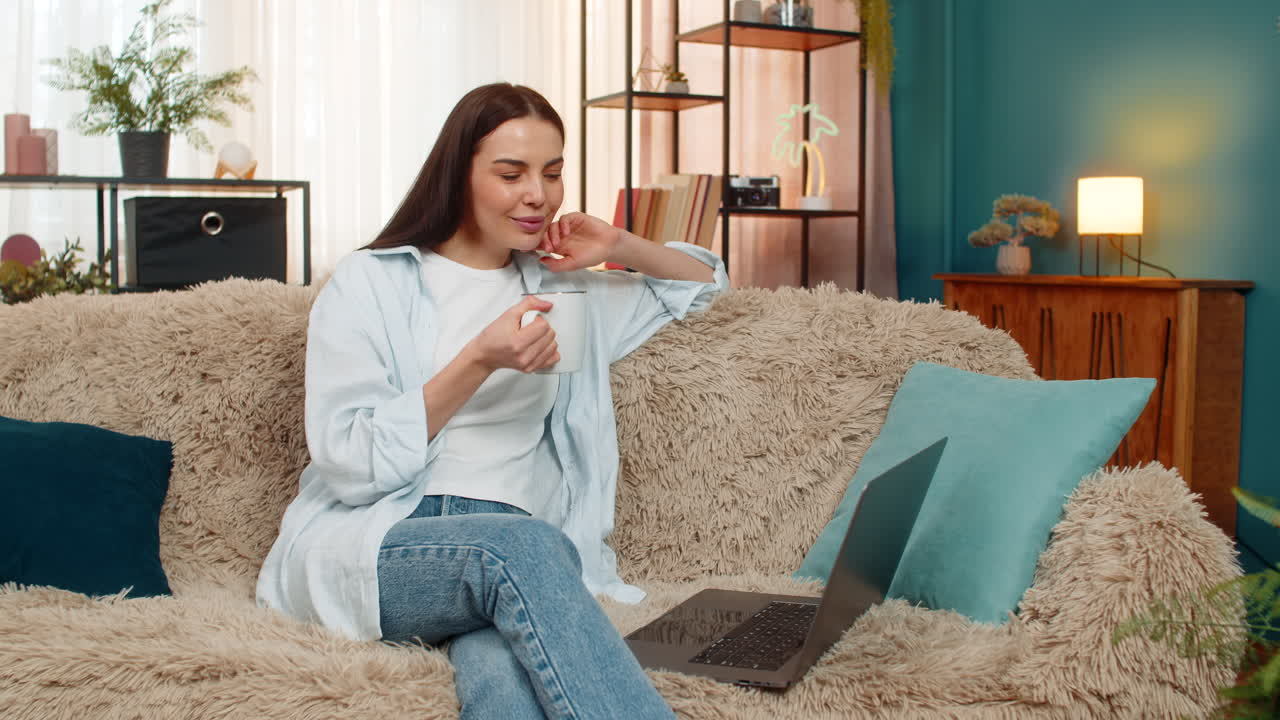 Young woman sitting on sofa happily watching online movie on laptop drinking tea relaxed enjoyment