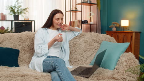 young woman sitting on sofa happily watching online movie on laptop drinking tea relaxed enjoyment