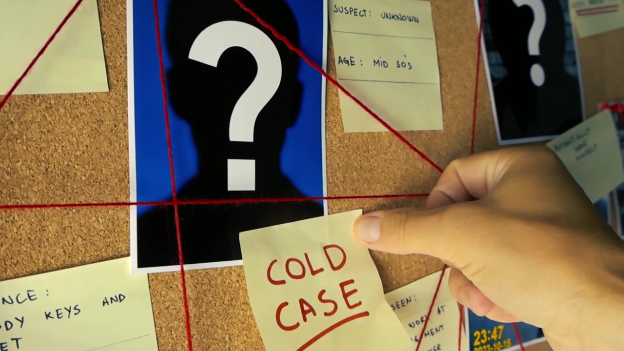 Investigator's hand is placing a yellow sticky note that says 'Cold Case' on a corkboard investigation map with photos, evidence notes and red string connecting the dots