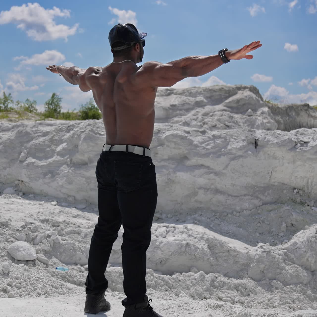 Muscular dark skinned man without shirt stands on white hill. Shirtless african athlete stands with stretched arms among summer nature. Sportsman in the mountains. Rear view