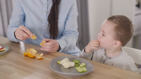 unrecognizable mom feeding her little son with clementine while sitting together at table in living room 2