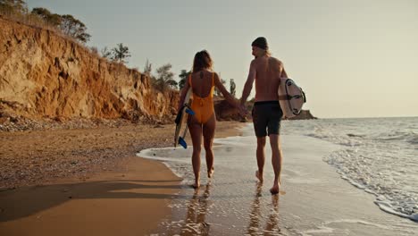 una pareja feliz, un chico rubio y su novia rubia en un traje de baño naranja, caminan y se toman de la mano a lo largo de la orilla rocosa de arena del mar, tienen tablas de surf en sus manos y caminan a lo longo de pequeñas olas en el mar por la mañana al amanecer