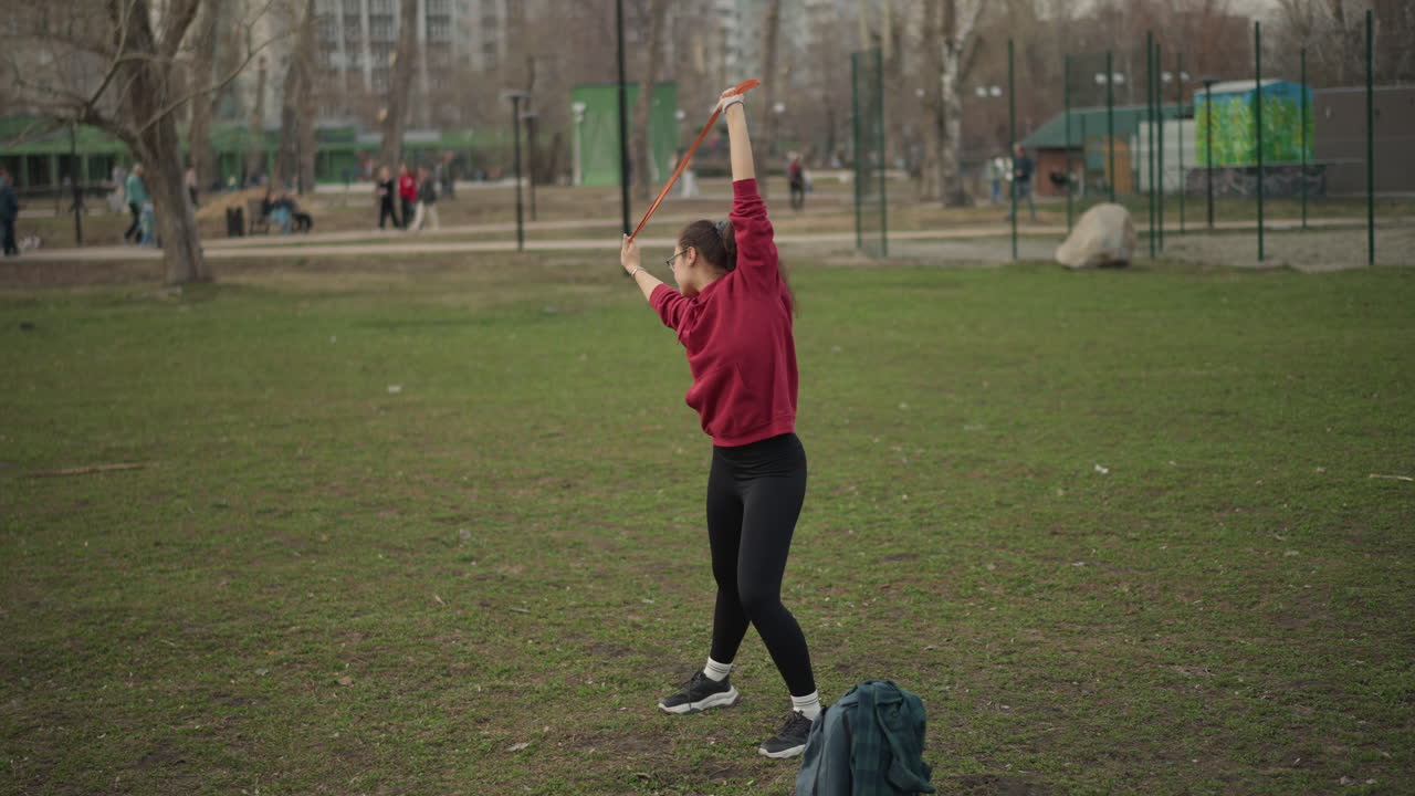 White Woman Practicing Flowing Hoop Choreography On Open Field With Fluid Arm Circles And Lunging Steps, Red Hoodie, Backpack At Side, Playground In Background, Expressive Athletic Movement