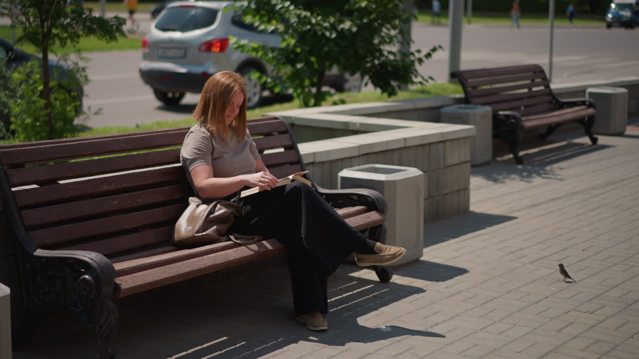 Student sitting on wooden bench reading book under bright sunlight, relaxed posture with handbag beside, background showing parked cars, trees, and blurred passersby
