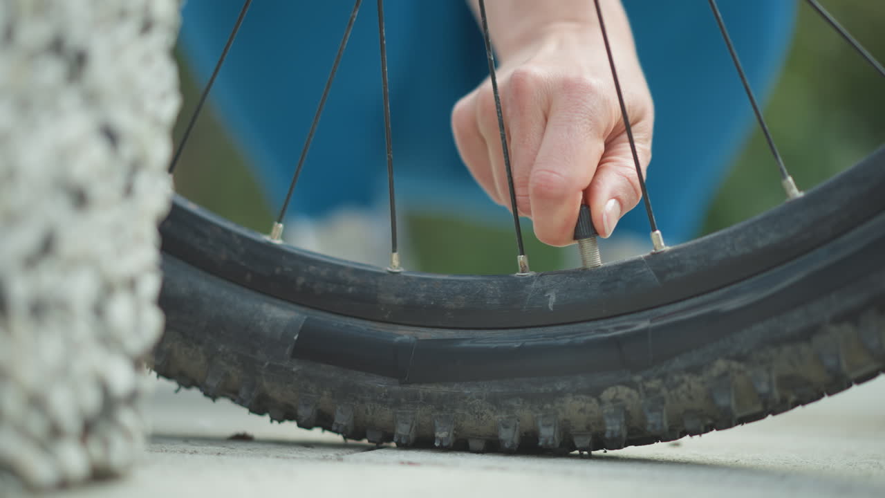 Primo piano della mano di un ciclista che estrae un chiodo incastrato nel pneumatico di una mountain bike con tassellatura grossa. La mano del ciclista impugna l'asta di metallo ed estrae il chiodo accanto a una giacca blu, con asfalto e fili d'erba. Soluzione rapida a bordo strada.