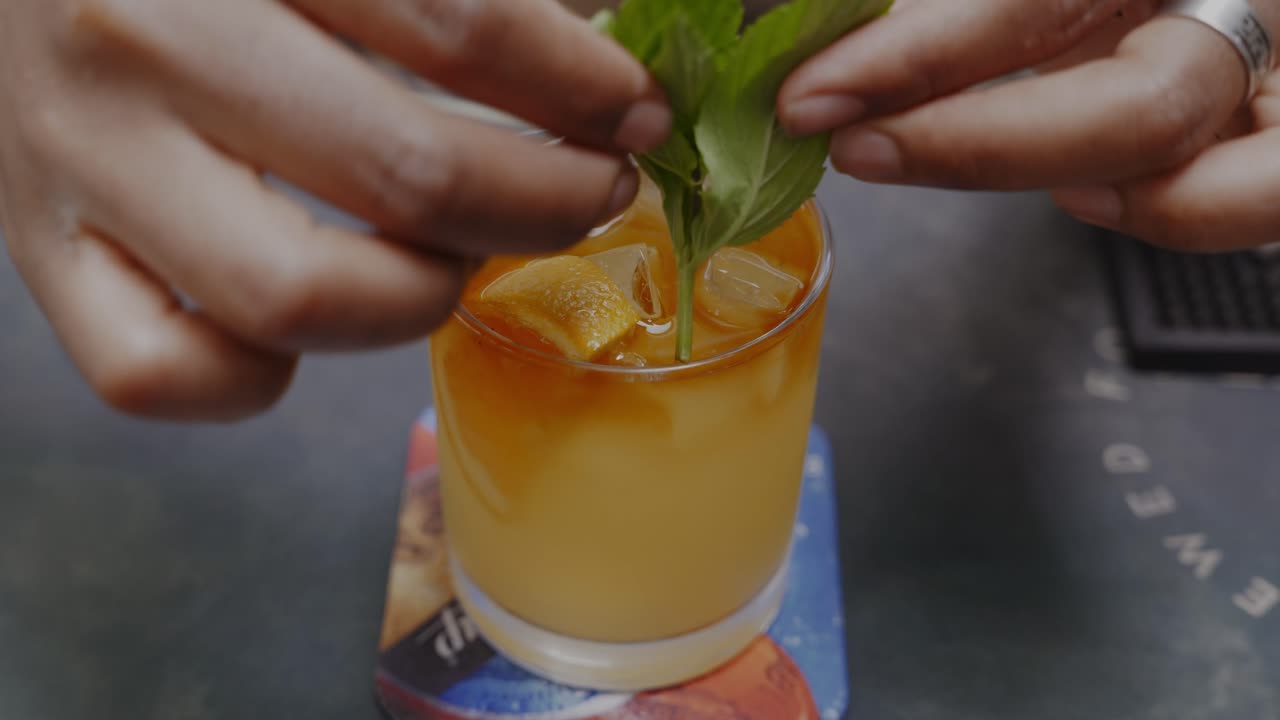 manos agregando menta y rodaja de naranja en jugo refrescante con cubitos de hielo