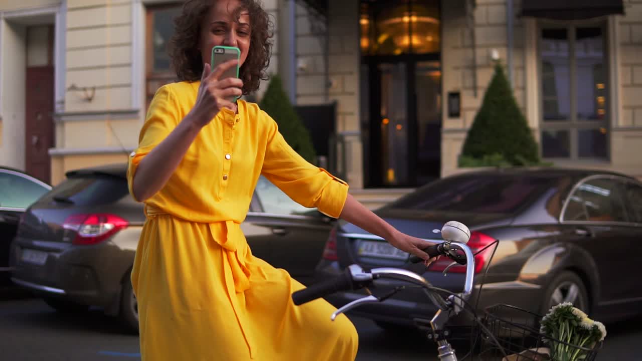 Beautiful smiling woman in long yellow dress riding a city bicycle with a basket and flowers inside and filming using her phone