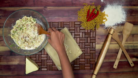 top view of making the spring rolls, marinated macaroni in the bowl, raw macaroni, wooden spoon, wooden spatula, flour, rolling pin, a frozen cheese piece and chili on the table