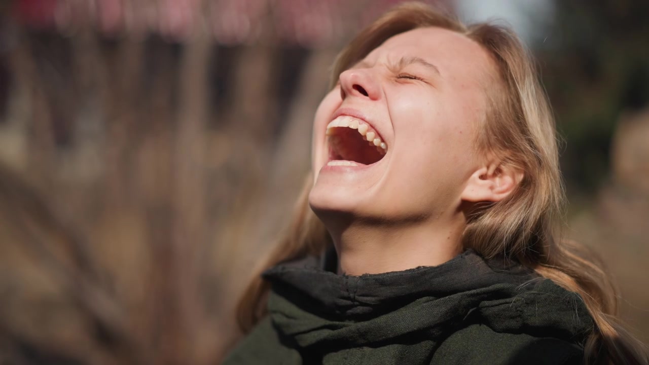 Ritratto a mezzo busto di giovane donna che ride con gli occhi chiusi, testa leggermente girata a destra e inclinata all'indietro, mostrando un sorriso a bocca aperta, guance animate e postura rilassata in una luminosa luce esterna.