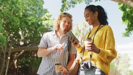 pareja feliz y diversa llamando a un taxi, usando un teléfono inteligente y sosteniendo un café para llevar en la ciudad