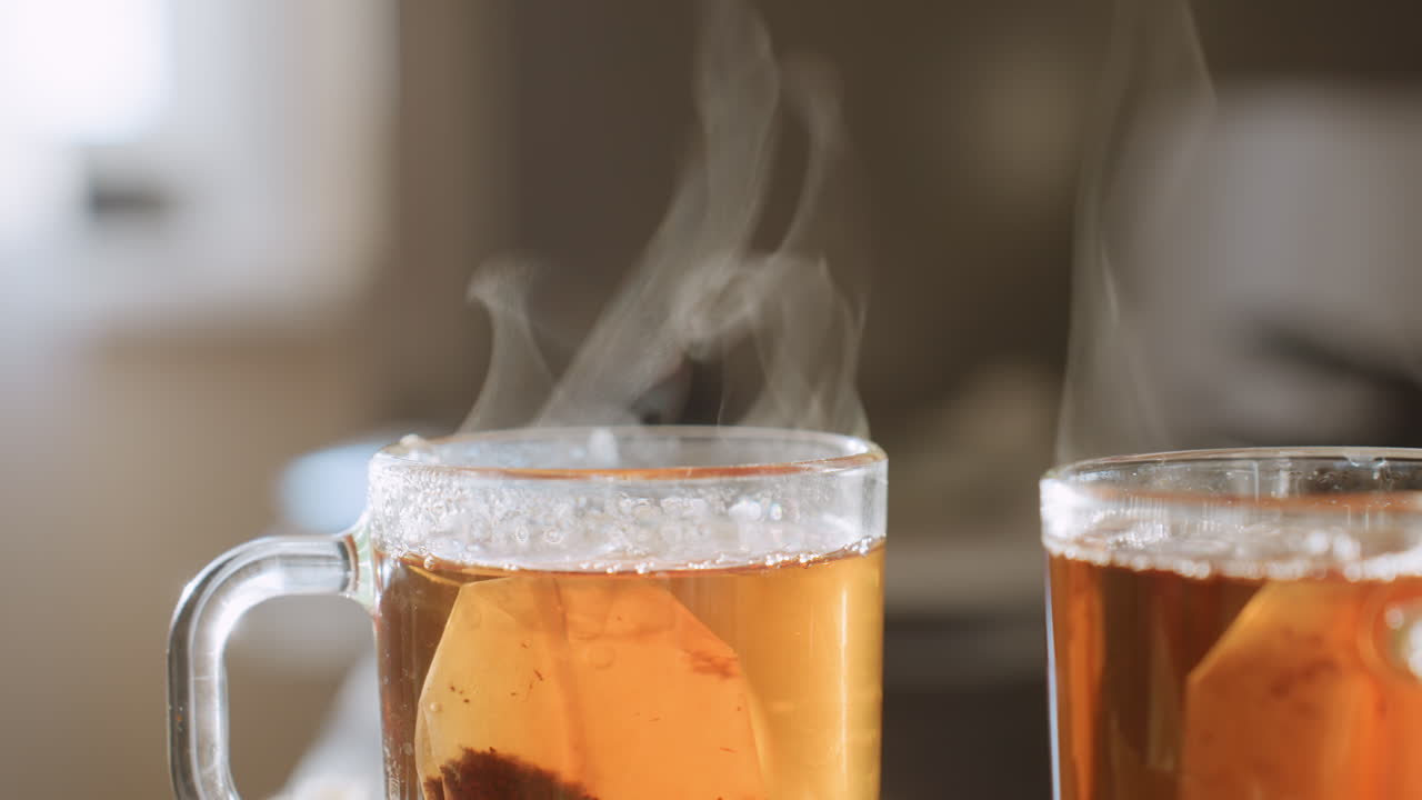 Close up of steaming herbal tea in transparent glass cup with teabag inside, vapor rising gracefully from hot surface, condensation forming around rim, warm amber liquid glowing in soft kitchen light