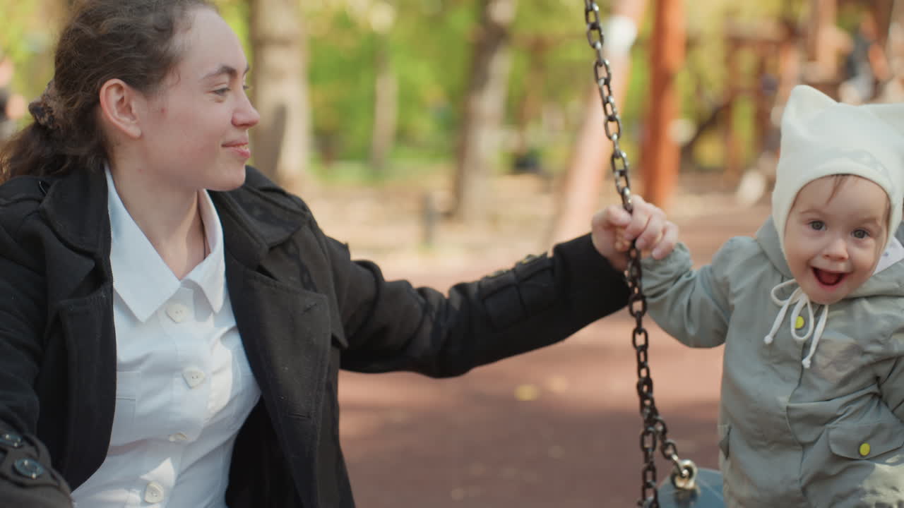 Criança ri alegremente. Criança jovem interage com a mãe ao ar livre. Bebê sorri brilhantemente sentado no balanço do parque com a mãe. Criança e mãe alegres desfrutam de atividades lúdicas ao ar livre juntas no parque