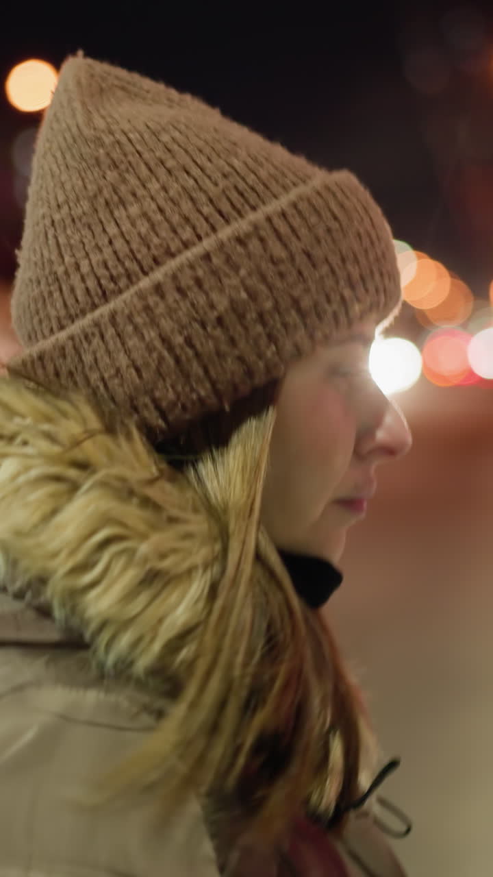 First-person view of a woman in a winter coat with a fur hood, crossing the road at night with blurry people and city lights in the background