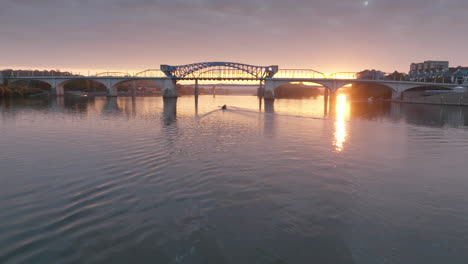 Flying-close-across-the-morning-water-of-the-Tennessee-with-boat-rowers-in-the-distance-silhouetted-by-the-sunrise-in-Chattanooga,-Tennessee