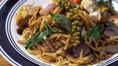 Tofu-Pad-Kee-Mao-Cubierto-Con-Albahaca-Tailandesa-Fresca-Y-Granos-De-Pimienta-Verde-En-Un-Plato-Azul-Y-Blanco-Sobre-La-Mesa,-Cierre-Deslizante-4k