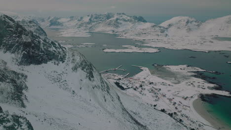 Aerial-view-across-peaceful-wintry-rocky-mountain-slopes-to-Ramberg-village,-Nordland,-Scandinavia