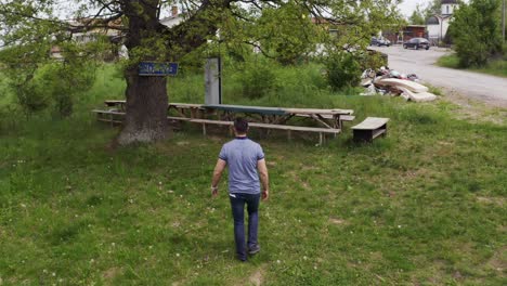 follow through drone shot following a tourist walking towards a tree and the benches located in the village of tsarichina hole in bulgaria