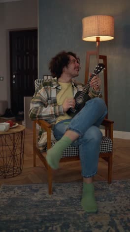 vertical video of a happy guy with curly hair in a plaid shirt sitting on a chair and playing a ukulele stringed musical instrument at home in the evening