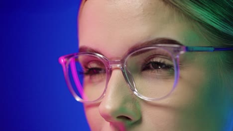 slow motion close up shot of a pretty attractive woman wearing glasses while she adjusts the glasses with her finger on her face with purple light in her face against blue background