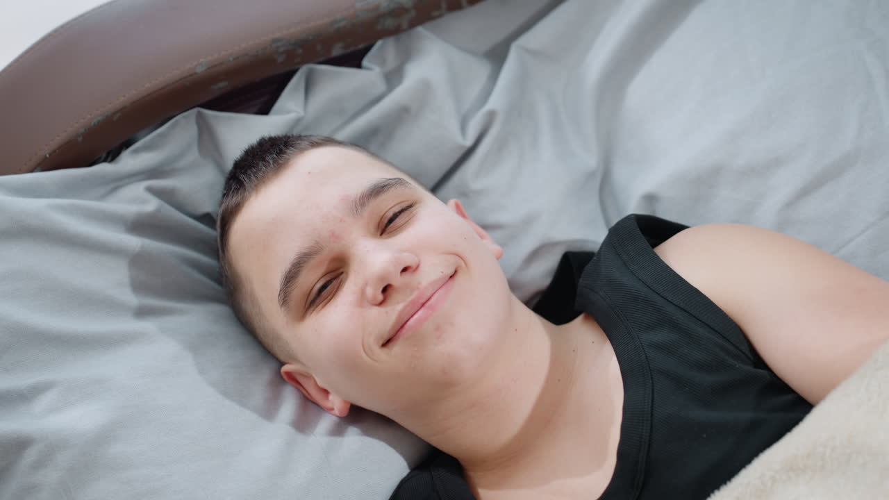 Top down view kid in black singlet peacefully sleeping on bed then slowly opens eyes and shows warm gentle smile under blanket creating tender atmosphere of comfort