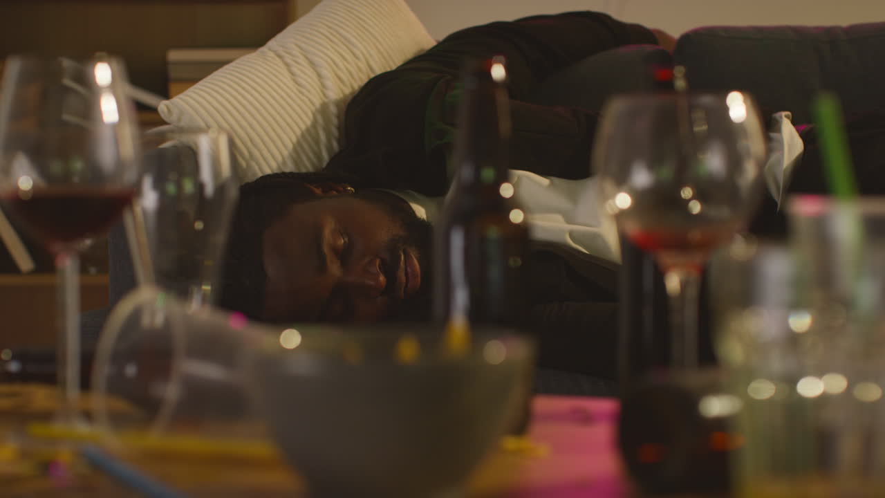 hombre tendido desmayado en el sofá después de beber alcohol en la fiesta de la casa sufriendo de resaca con botellas vacías y vasos en primer plano