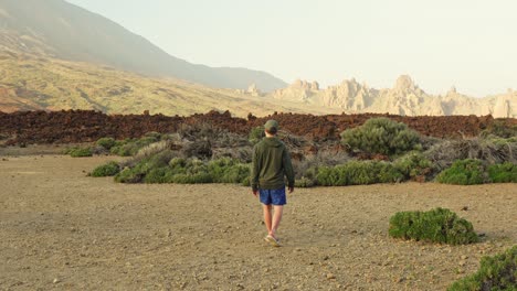 man walking alone across the land towards the mountains while wearing jacket hoodie and baseball cap, static wide angle