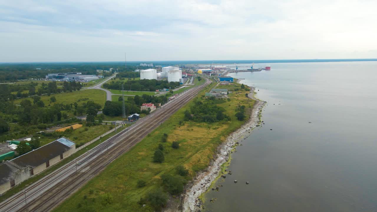 Aerial drone footage flying above a seaside industrial area with a railroad beneath and a rocky beach shoreline on the side. Trading port with cranes and liquid storage white container vessels visible