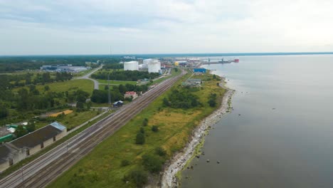 aerial drone footage flying above a seaside industrial area with a railroad beneath and a rocky beach shoreline on the side. trading port with cranes and liquid storage white container vessels visible