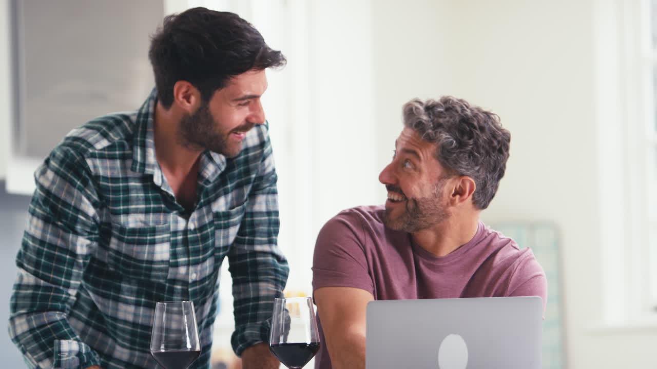 동성 남성 커플이 와인을 마시고 집에서 노트북을 사용하여 재정을 확인하고 휴가를 예약하거나 쇼핑합니다.