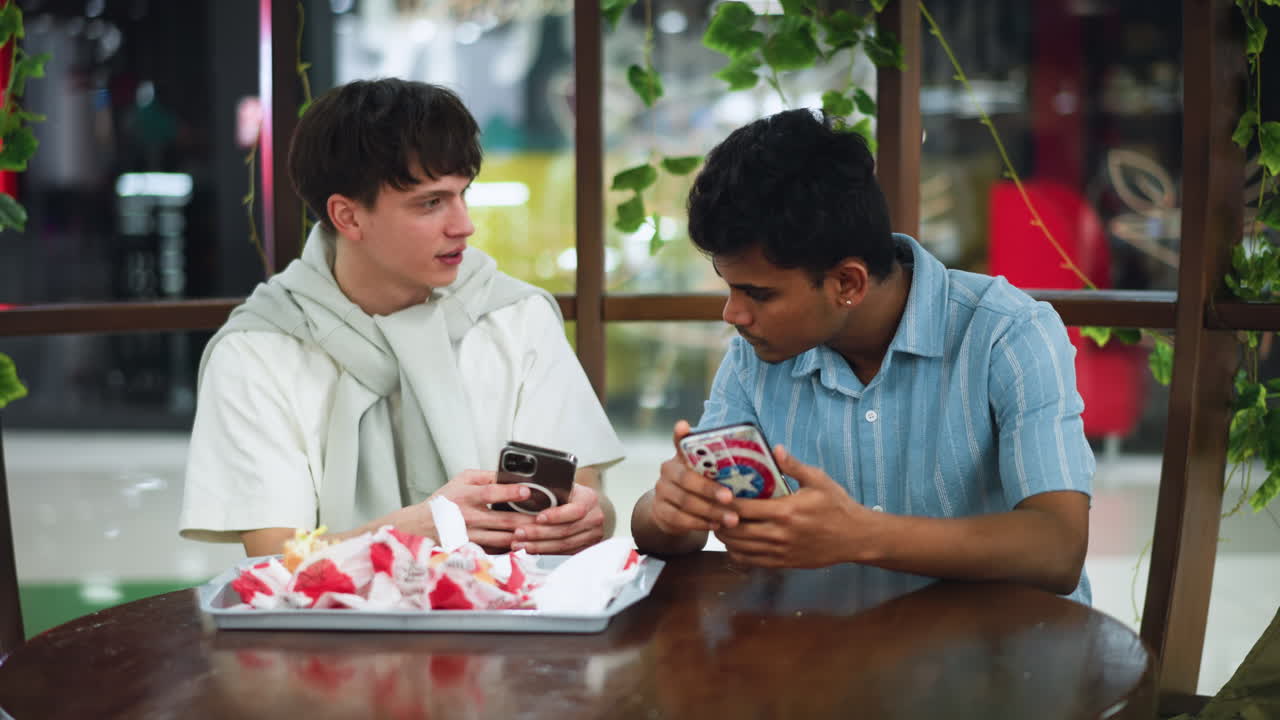 Two best friends analyzing phone while sharing burger and chips on white tray during casual conversation, close shot capturing focused expressions and snack items, relaxed indoor hangout moment