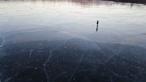 man walking alone on thick ice surface of frozen lake in twilight glow at sunset