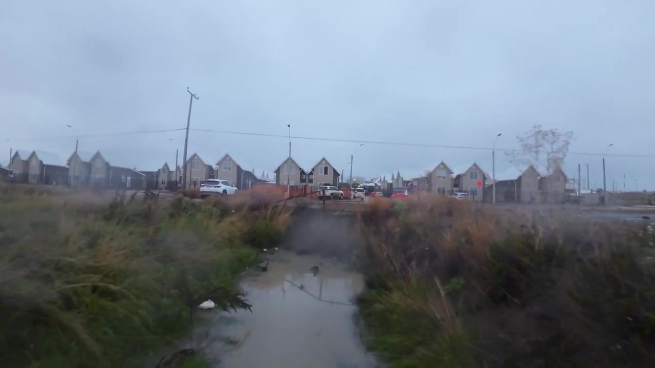 A fjord creek cuts through golden windswept grass toward distant houses near Puerto Natales, Chile under a misty overcast sky, low altitude FPV drone flight