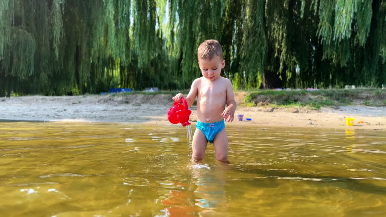 Cute Caucasian toddler boy stands in the river. Beautiful baby in swimming trunks fills his can and pours water.