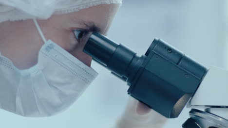 close-up of female scientist in protective mask looking in microscope