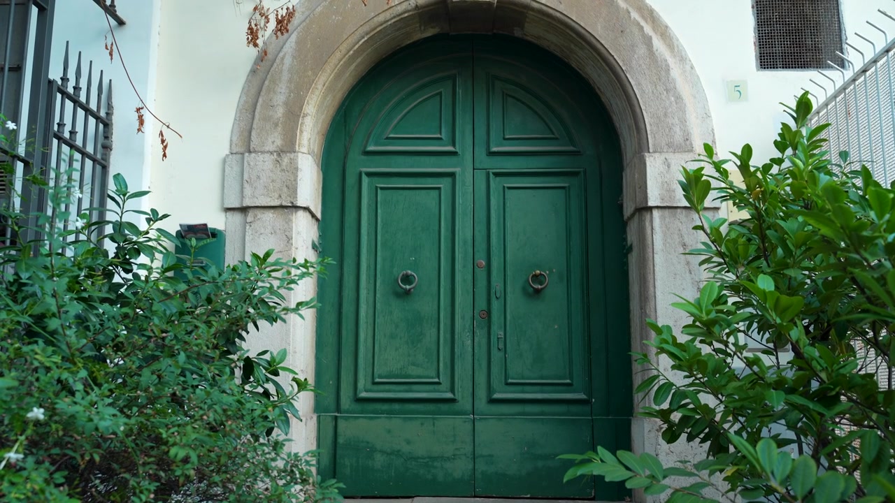 Green double wooden doors under a stone arch are framed by dense shrubs and railings in a narrow Vietri Italy doorway scene.