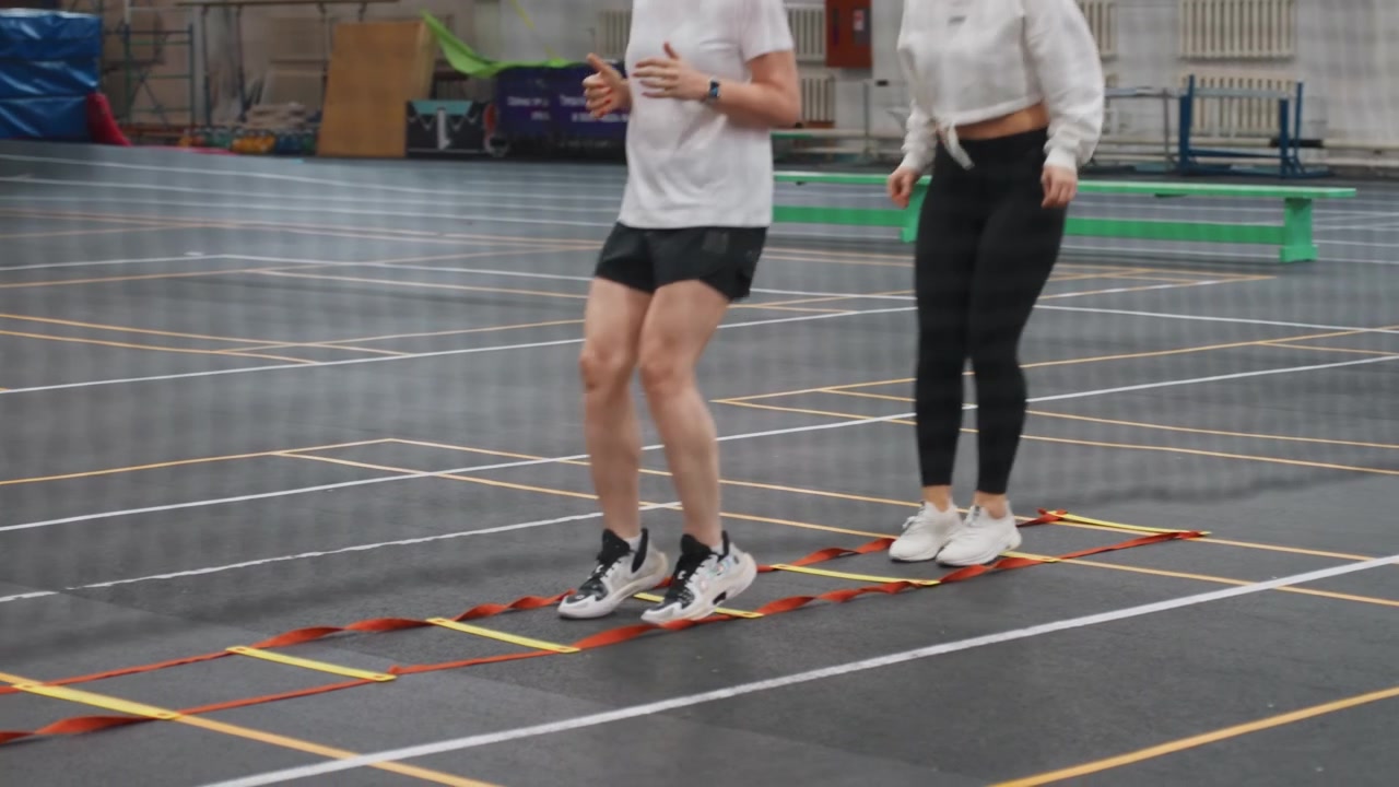Two caucasian women practicing badminton footwork during rehabilitation session on indoor court, physiotherapist guiding controlled jumps, focus on mobility, stability, gradual recovery and measured