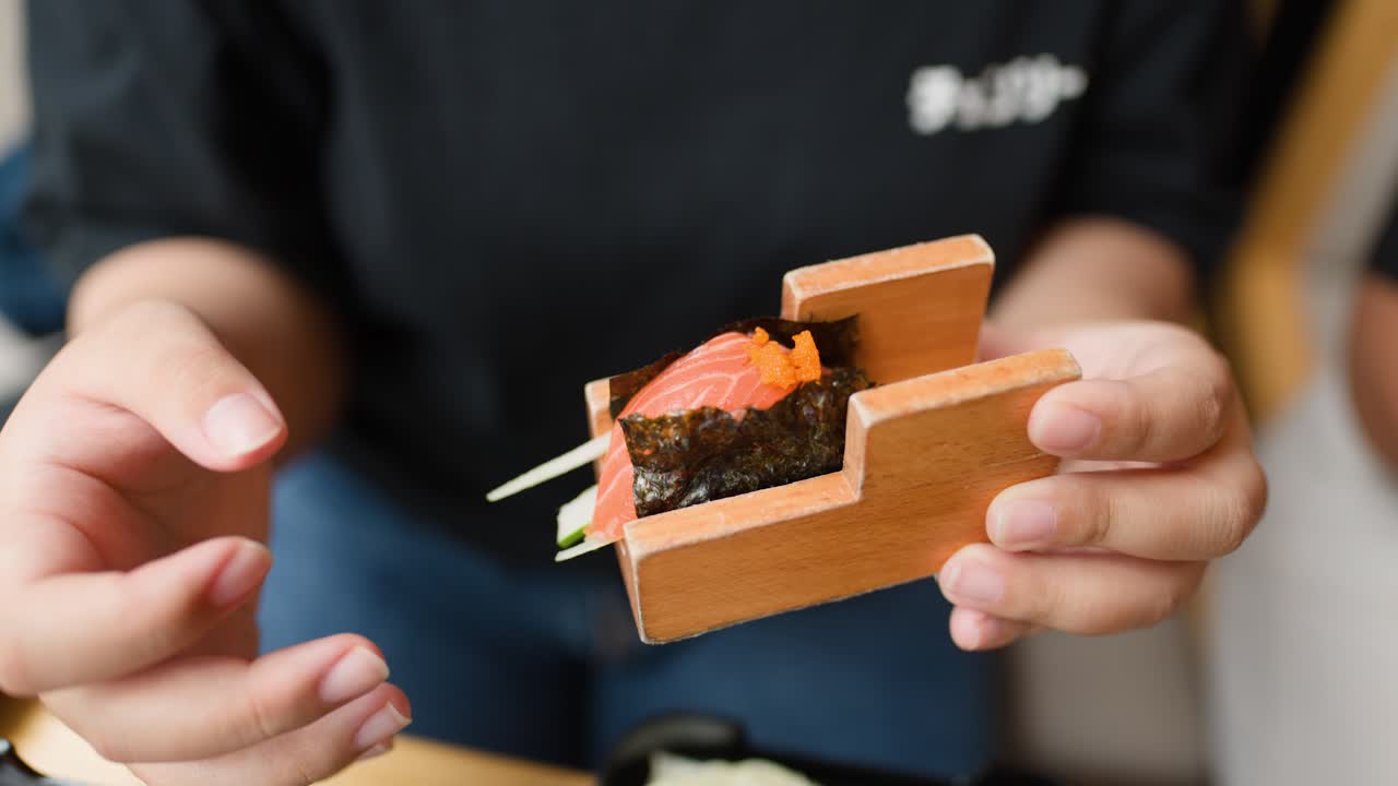 Person lifts fresh salmon sushi hand roll from wooden holder in bright, casual restaurant setting