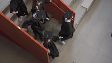 vista superior de un grupo de estudiantes de preescolar con toga y birrete corriendo por las escaleras sosteniendo sus diplomas de graduación mientras uno de ellos abraza al maestro