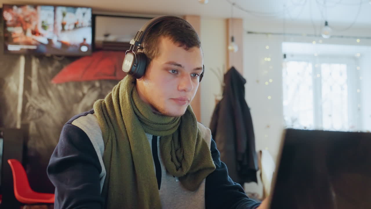 Caucasian man at laptop wearing headphones, focused on screen while tuning headset and typing, holiday lights and window light in background, cafe interior with pastry counter, serious expression