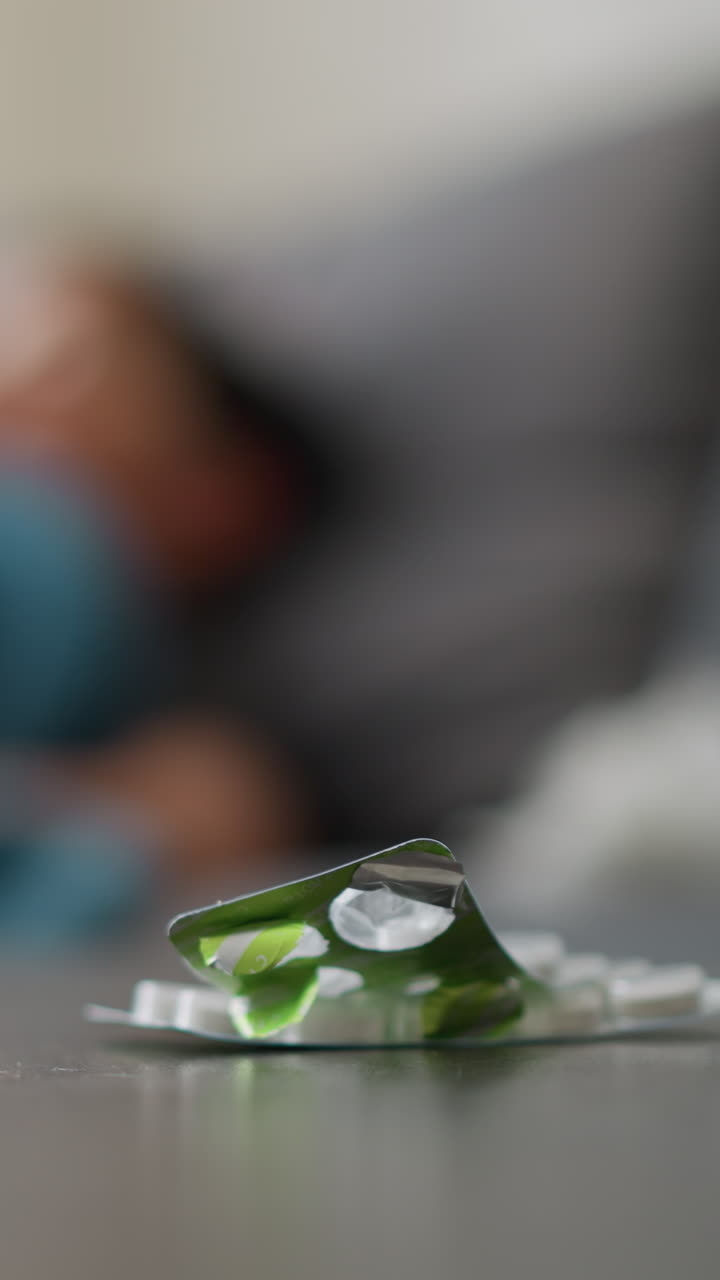Close-up of medication and cup on table with blurred view of person adjusting on bed, suggestive of recovery, rest, and care during illness with soothing indoor setting